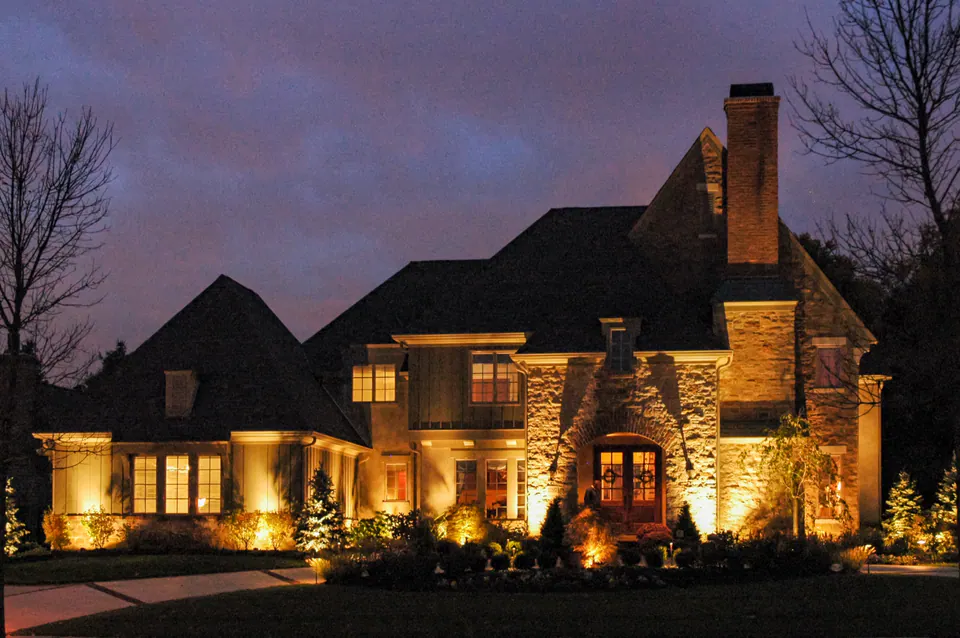 Stone home facade, front entry, and foundation planting with warm architectural uplighting at dusk